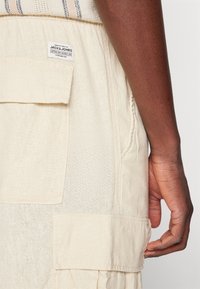 Beige bomullsplagg med en texturerad yta, med en ficka i lappar, sydda detaljer och en etikett på baksidan.