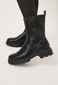 Botas de cuero negras hasta el tobillo con cremallera lateral, suela de goma gruesa y sutiles detalles de costura. La textura es suave y pulida.
