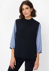 Dunkelblauer, gerippter Strickpullover mit hohem Kragen, getragen über einer hellblauen Bluse mit langen Ärmeln. Besitzt einen lässigen Schnitt und subtile seitliche Akzente.