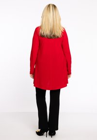 Rode lange mouwen tuniek met een vloeiende uitstraling, gezien van achteren, gecombineerd met zwarte strakke broek en zwarte hoge hakken.
