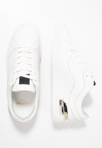 White leather sneakers feature textured accents, round toe, flat sole, and metallic heel detail. Laces and black logo patch on the tongue.