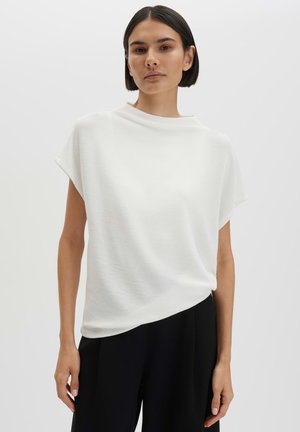 Vrouw met kort donker haar, gekleed in een losse witte top met korte mouwen en zwarte broek, staand tegen een effen lichte achtergrond.