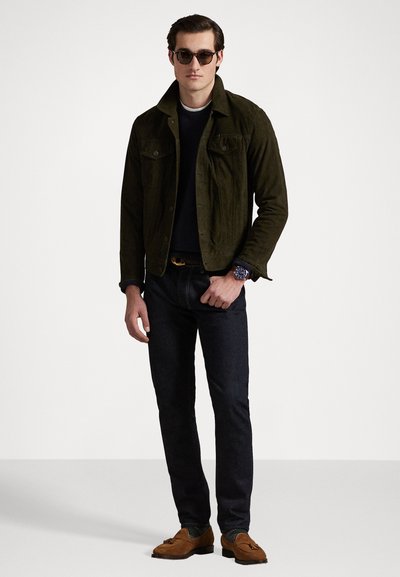 Chaqueta de ante verde, suéter negro, jeans oscuros y zapatos de ante marrón. Gafas de sol redondeadas y reloj de pulsera completan el atuendo.