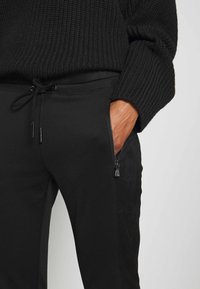 Svarta joggarbyxor med dragsko i midjan, med en dragkedjeficka på sidan. Materialet verkar mjukt med en slät textur och en minimalistisk design.