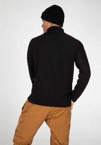 Maglione in pile nero a maniche lunghe con colletto alto, abbinato a pantaloni cargo color sabbia con tasche laterali. Berretto di maglia nero a forma circolare.