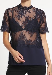 Blusa blu scuro con sovrapposizione in pizzo trasparente con motivi floreali, maniche corte e pannello inferiore solido. La rifinitura in pizzo aggiunge dettagli di consistenza.
