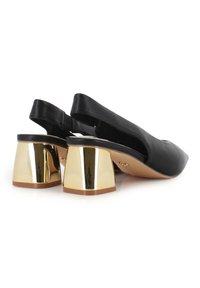Zapatos de cuero negro de tipo slingback con punta afilada, que presentan un tacón cuadrado de bloque metálico dorado para añadir contraste y estabilidad.