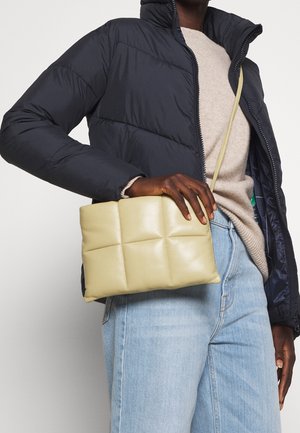 Personne portant une doudoune noire, un pull beige et un jean bleu clair, tenant un sac bandoulière matelassé beige au niveau de la taille.