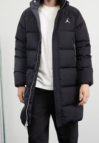 Zwarte pufferjas met ritssluiting aan de voorkant, met grote gewatteerde secties, een hoge kraag en een wit Jordan-logo op de linkerkant van de borst.