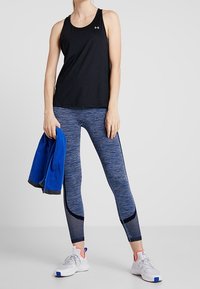 Czarny sportowy top na ramiączkach, niebieskie legginsy w wzory z siateczkowymi akcentami oraz szare sneakersy z różowymi detalami, trzymająca niebieską kurtkę.