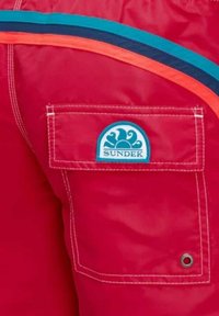 Pantaloni corti impermeabili rossi con tasca laterale, caratterizzati da un logo "SUNDEK" blu e bianco e strisce arancioni e blu scuro lungo la vita.