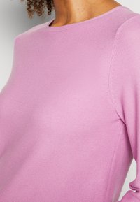 Gros plan d'une personne portant un pull en tricot violet clair à col rond et manches longues, mettant en évidence la texture du tissu et la couture de l'épaule.