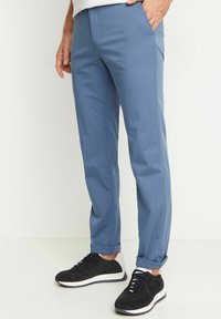 Pánské modré chinos kalhoty se slim fit střihem, plochým předním dílem a bočními kapsami. Nosí se s černými teniskami se strukturovanou textilií a bílými podrážkami.