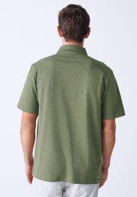 Polo majica kratkih rukava u maslinasto zelenoj boji, s klasičnim ovratnikom i teksturiranim pletenim materijalom. Jednostavan dizajn s ravnim rubom na stražnjoj strani.