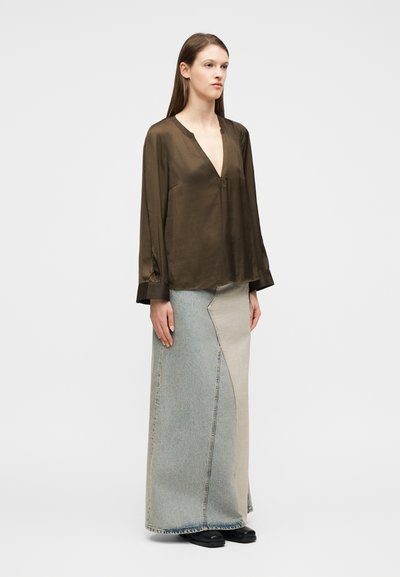 Γυναίκα που φοράει σκούρο καφέ μπλούζα με μακριά μανίκια και βαθύ V-λαιμόκοψη, μαζί με  μια δίχρωμη φούστα από ανοιχτό και μεσαίο ντένιμ, στέκεται σε λευκό φόντο.