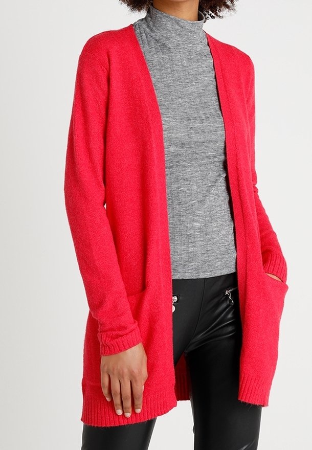Femme portant un cardigan rouge vif ouvert sur le devant, sur un haut gris à col montant et un pantalon noir avec des détails zippés.