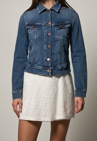 Blå jeansjacka med kopparfärgade knappar, två bröstfickor och en kortare design. Bärs över en vit texturerad kjol.