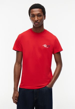 Mężczyzna w czerwonej koszulce Tommy Jeans z białym i granatowym logo, ciemnoniebieskie dżinsy, stoi z jedną ręką w kieszeni na białym tle.