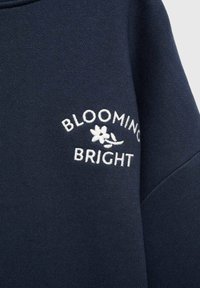 Mörkblå sweatshirt med vit broderad text "BLOOMING BRIGHT" och en blommig accent, tillverkad av mjukt, texturerat tyg.