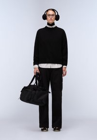 Il modello indossa un maglione di lana nero, sovrapposto a una camicia bianca a righe. Pantaloni cargo neri, una grande borsa a tracolla nera e cuffie rotonde nere.