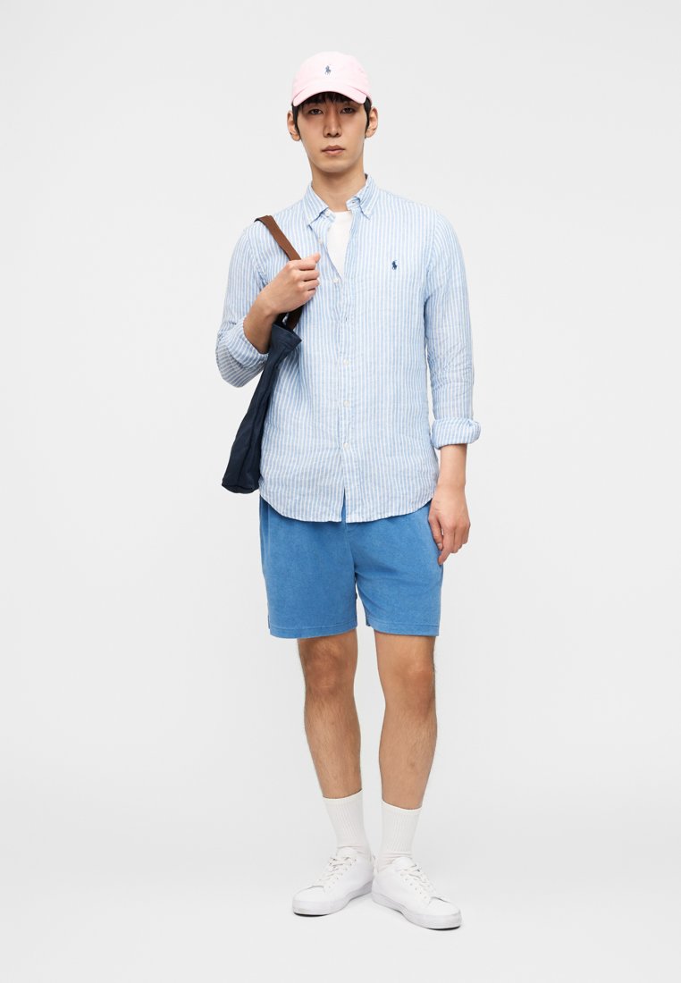 Young man wearing a pink cap, blue striped button-up shirt, blue shorts, white socks and sneakers, holding a navy shoulder bag.