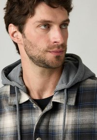 Giacca a camicia a quadri con cappuccio grigio, colori nero, bianco e blu, chiusura con bottoni e design del tessuto testurizzato.