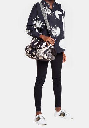 Personne portant une chemise noire à motifs, un pantalon skinny noir, des baskets blanches avec des rayures décoratives, tenant un sac à bandoulière noir et blanc à motifs.