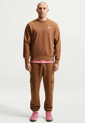 Hombre con la cabeza afeitada, lleva una sudadera marrón de Nike y pantalones cargo, camiseta interior rosa y zapatillas rosas, de pie y mirando hacia adelante sobre fondo blanco.