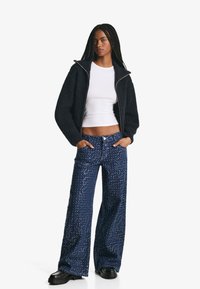 Námornícky textúrovaný zipsový kabát cez biele crop top, spárovaný s širokými nohavicami so flitrami. Čierne robustné topánky dotvárajú outfit.