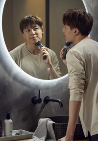 Junger Mann benutzt elektrischen Rasierer und schaut in einen runden, beleuchteten Badezimmerspiegel über einem Waschbecken mit schwarzem Wasserhahn.