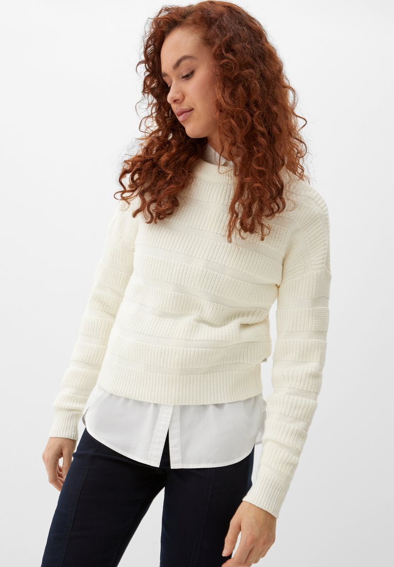 Ivory gebreide trui met gestructureerde horizontale strepen, ronde halslijn en nauwsluitende manchetten, gedragen over een wit shirt, gestyled met donkere broeken.