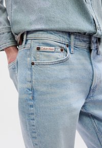 Ανοιχτό μπλε τζιν με κλασικό σχέδιο πέντε τσεπών, με μπρούτζινα εξαρτήματα και ετικέτα με το λογότυπο Calvin Klein στην πίσω τσέπη.