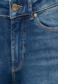 Gros plan d’un jean en denim bleu montrant le bouton avant, la zone de la fermeture éclair et la poche avec des détails de couture orange.