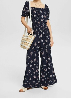 Frau trägt marineblauen Jumpsuit mit Blumenmuster und Puffärmeln, hält eine geflochtene Korbtasche und trägt weiße Sandalen.