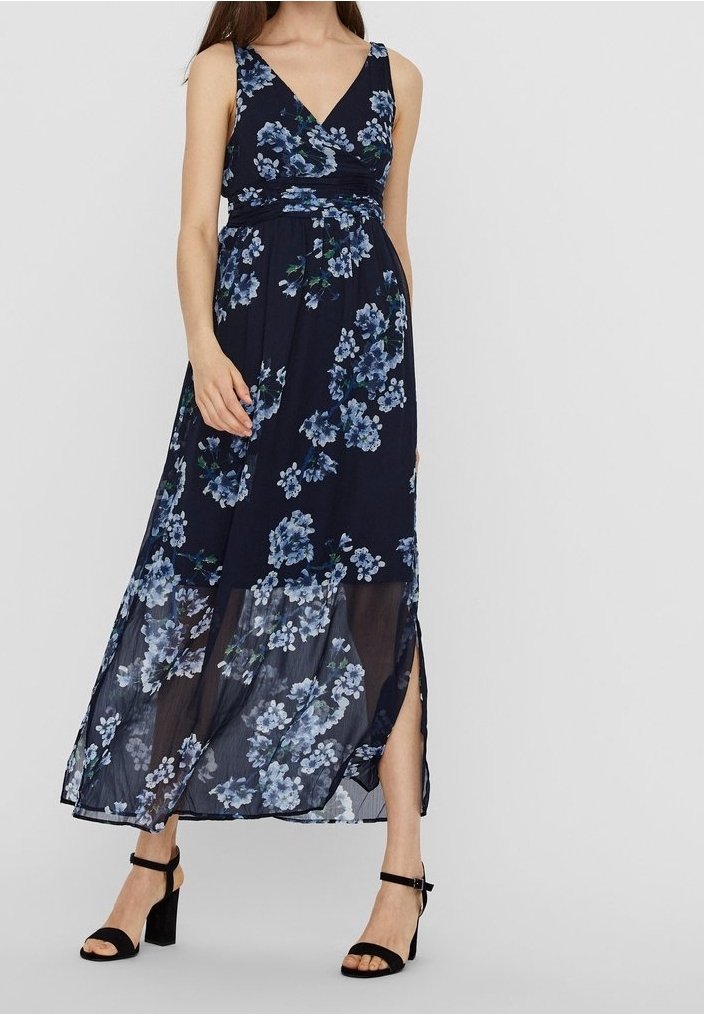 Femme portant une robe longue sans manches bleu marine avec des motifs floraux bleus et une fente latérale, assortie à des sandales à talons noires avec bride à la cheville.
