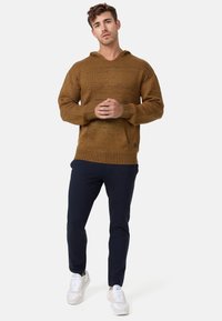 Hoodie en tricot marron avec une finition texturée, grande poche avant, poignets côtelés, associé à un pantalon slim-fit bleu marine et des baskets blanches.