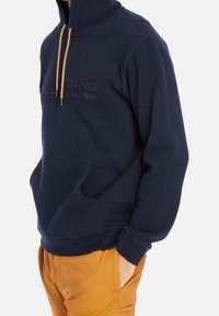 Marineblauwe fleece hoodie met hoge kraag, buidelzak en reliëflogopatch. Voorzien van gouden trekkoordaccenten en ribbelmanchetten.