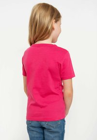 Child with long light brown hair wearing a bright pink short-sleeve shirt and blue jeans, facing away against a plain white background.