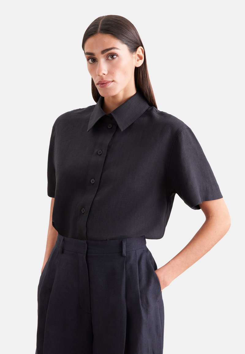 Femme aux cheveux raides et foncés portant une chemise noire à manches courtes boutonnée, rentrée dans un pantalon noir taille haute, les mains dans les poches.