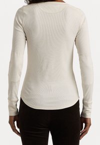 Długi rękaw, kremowy top wykonany z prążkowanej tkaniny; posiada łaty na łokciach oraz zaokrąglony dół.