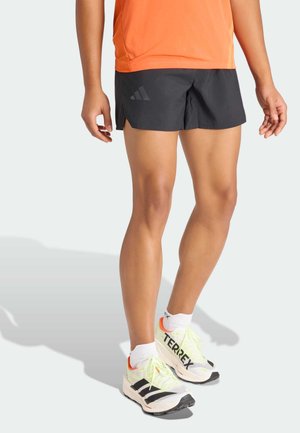 Shorts noirs avec fentes latérales, chemise en mesh orange et chaussures de course blanches avec des accents noirs et néon. Logo Adidas sur les shorts.