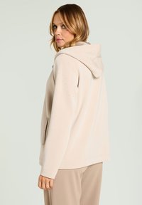 Veste à capuche beige en tissu doux, avec de petites perforations, des poches latérales et une coupe décontractée, portée avec un pantalon beige.