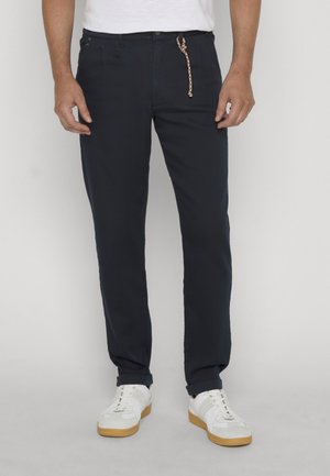 Homme portant un pantalon bleu marine avec un cordon tressé et des baskets blanches, debout devant un fond uni.