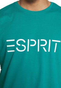 Tyrkysové bavlněné tričko s bílým logem se stylizovaným nápisem "ESPRIT" tištěným na hrudi. Krátké rukávy, kulatý výstřih.