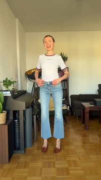 Νεαρή γυναίκα που χαμογελά, στέκεται σε σαλόνι φορώντας λευκό μπλουζάκι, ανοικτό μπλε τζιν και καφέ παπούτσια, με ορατά τατουάζ στα χέρια της.