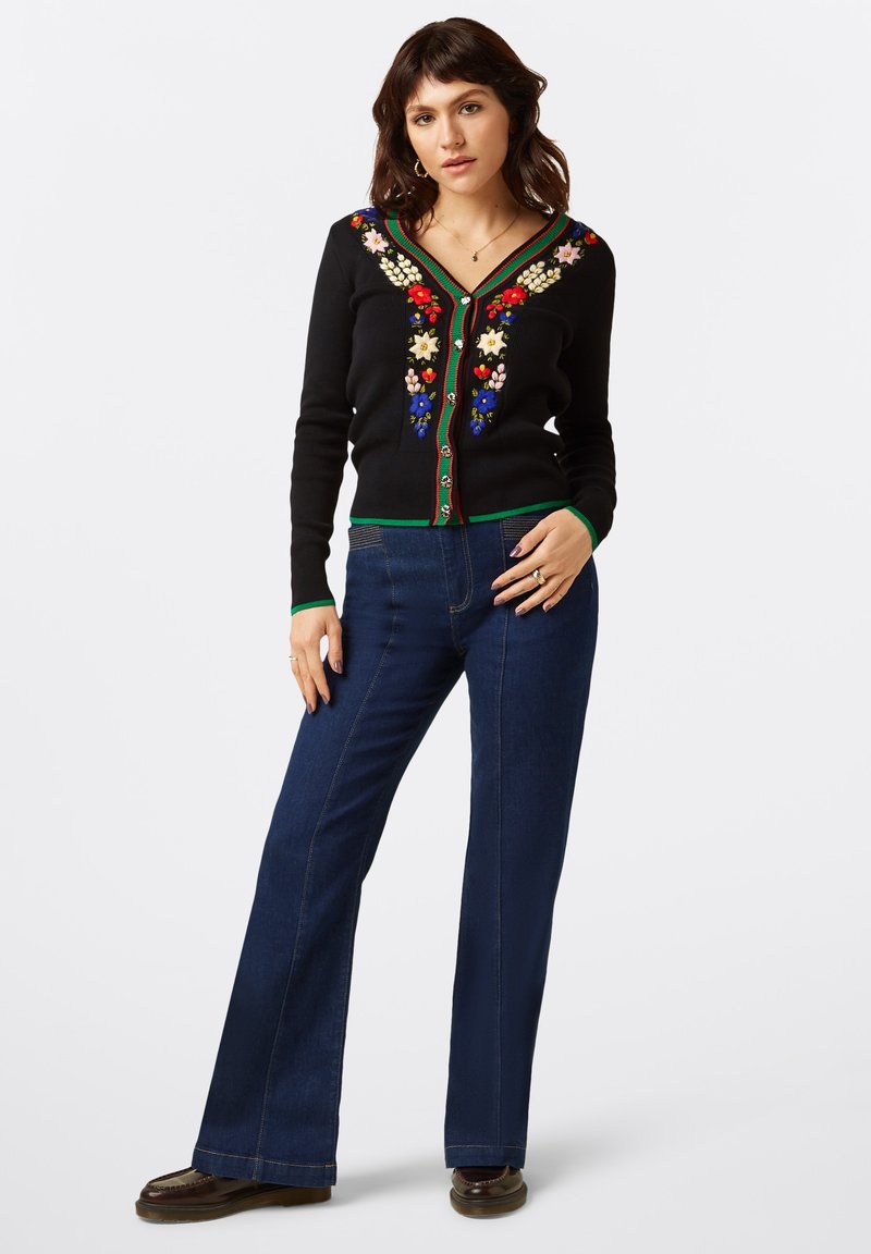 Cardigan noir avec une broderie florale colorée le long du col et sur le devant, doté d'une fermeture à boutons et d'accents verts. Associé à un jean bleue évasé.