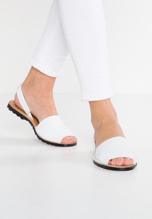 Stopy w białych sandałach typu slingback z odkrytymi palcami i czarnymi podeszwami, w zestawieniu z białymi dopasowanymi spodniami na jednolitym białym tle.
