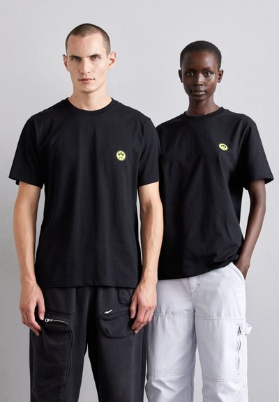 T-shirts en coton noir à manches courtes, arborant de petits logos de smiley néon sur la poitrine. Portés avec des pantalons cargo de couleurs foncées et claires.