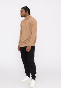 Pull à col roulé tricoté beige avec un motif texturé, associé à un pantalon cargo noir avec poches latérales et des baskets blanches.