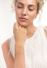 Bracelet en métal doré porté au poignet, avec deux bagues fines. Le modèle a des cheveux bouclés et porte un haut blanc texturé avec des franges.